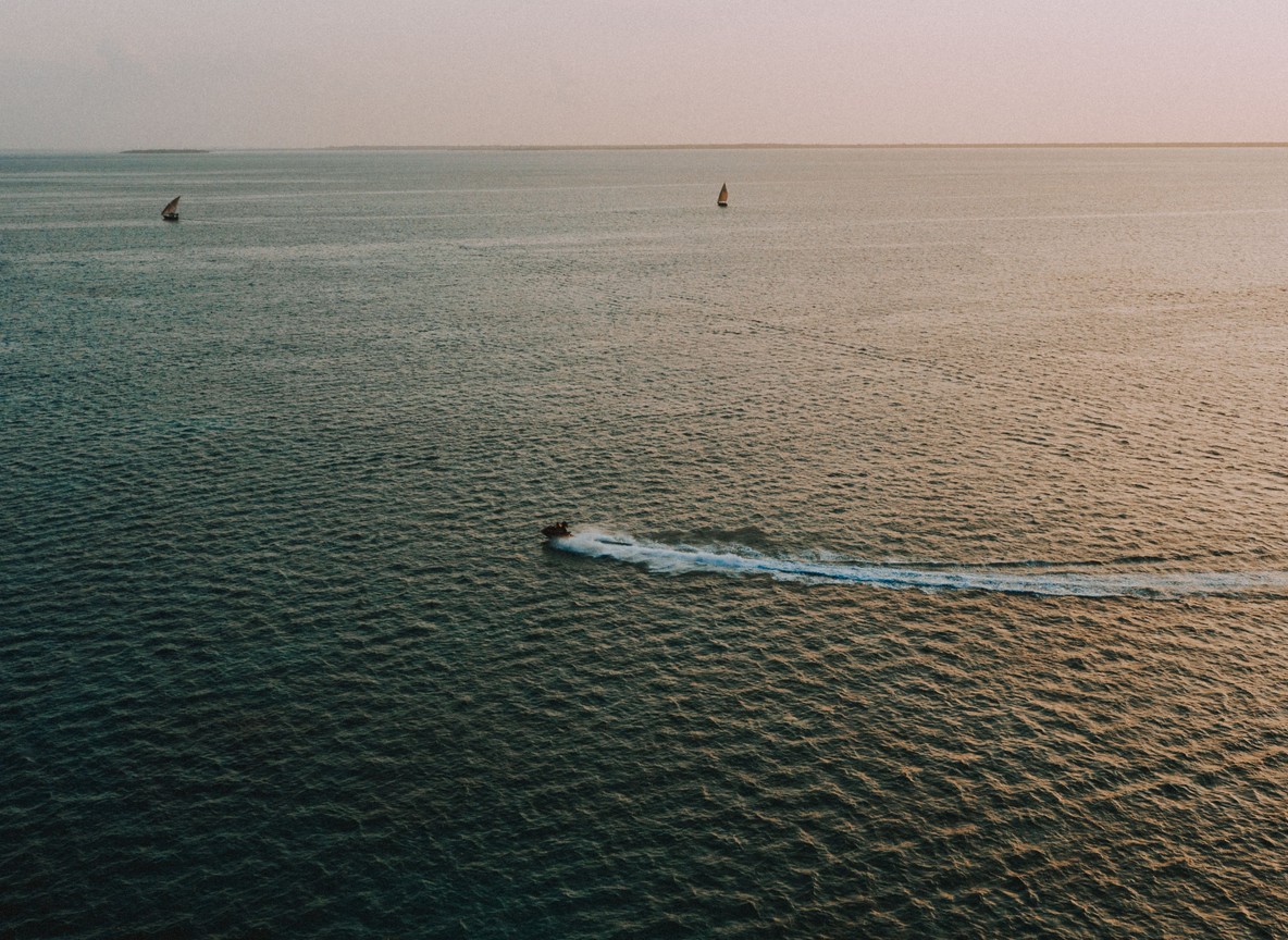 Traditional dhow sailboat with white sail crossing the channel between Lamu and Manda Island at sunset