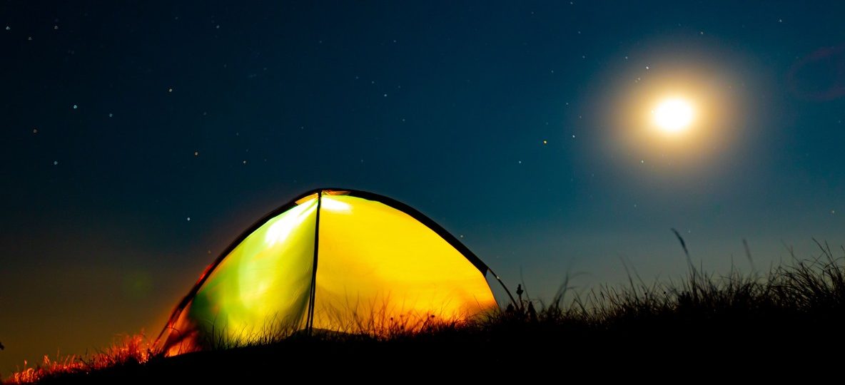 An open-air safari tent glowing at night under a star-filled sky and rising moon in the Kenya bush